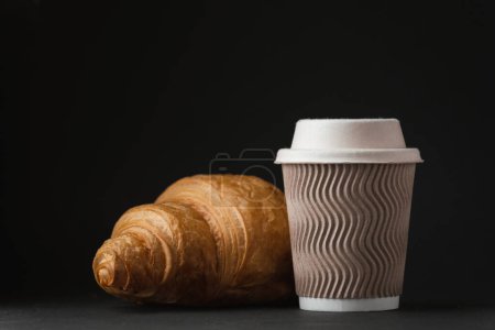 Paper cup of hot coffee with paper lid next to freshly baked croissant on the black background. Mock up with copy space for a free text