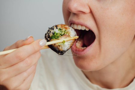 Téléchargez les photos : Femme apporte rouleau de sushi en utilisant des baguettes à sa bouche et aime la nourriture japonaise - en image libre de droit