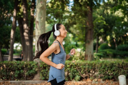 Photo for Happy asian athlete girl running in the park while she is listening to music on white headphones - Royalty Free Image