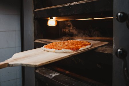 Téléchargez les photos : Baker sortant une pizza chaude du four avec une pelle en bois dans une boulangerie - en image libre de droit