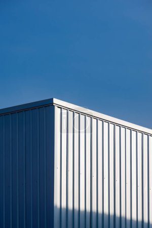Foto de Pared del edificio del almacén de acero corrugado con la reflexión de la luz del sol en superficie contra fondo azul claro del cielo en marco vertical - Imagen libre de derechos
