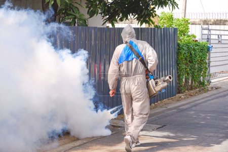Téléchargez les photos : Vue arrière du travailleur de la santé dans les vêtements de protection à l'aide d'une machine à brouillard pulvérisation chimique pour éliminer les moustiques dans la rue dans l'allée de la communauté publique de banlieue - en image libre de droit