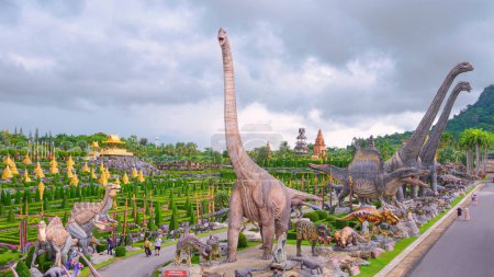 Foto de Chonburi, Tailandia - 24 de septiembre de 2023: Grupo de grandes estatuas de dinosaurios y hermoso jardín topiario con turistas caminando por la pasarela en el maravilloso valle tropical del mundo en el jardín Nong Nooch, Pattaya - Imagen libre de derechos