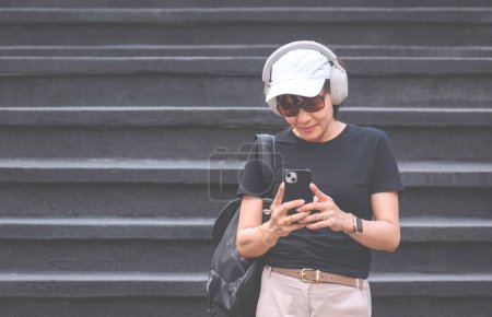Téléchargez les photos : Une touriste asiatique dans un style vestimentaire décontracté prenant selfie ou vidéo avec téléphone portable dans la zone d'attraction extérieure tout en créant du contenu de voyage sur les médias sociaux. - en image libre de droit