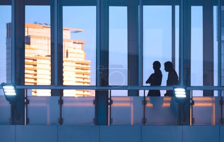 Foto de Silueta de 2 turistas femeninas caminando por la pasarela de cristal elevada del edificio moderno con fondo borroso de reflejo de la luz del sol dorada en el rascacielos en el momento de la puesta del sol - Imagen libre de derechos