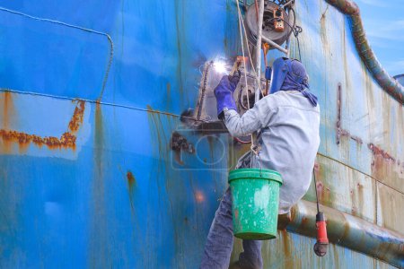 Photo for Welder rappelling with ladder to welding metal hull surface of the old fishing boat during maintenance and improvement work in shipyard area - Royalty Free Image