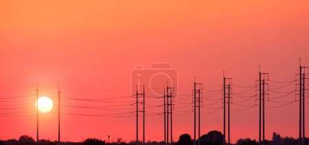 Fila de siluetas de postes eléctricos con líneas de cable contra fondo naranja del cielo al atardecer en el campo, vista panorámica con espacio para copiar