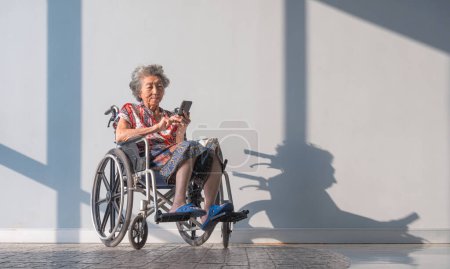 Asian mental healthy elderly woman using mobile phone on wheelchair relaxing in rest area while waiting for her annual medical check up inside of hospital. Health care and good mental health concept