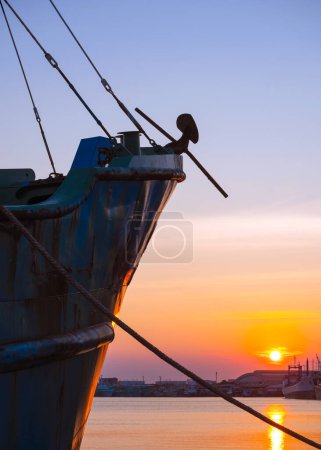 Silhouette forward side of fishing boat moored at harbor with mooring rope against sunset sky background in evening time and vertical frame