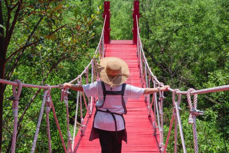 Rückansicht einer Touristin, die über eine rote hängende Holzbrücke vor grünem Hintergrund im Mangrovenwald im Naturpark spaziert, Ökotourismus-Konzept