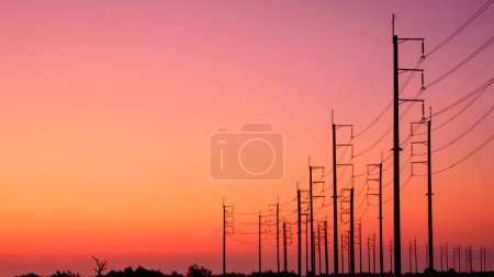 Silueta dos filas de postes eléctricos con líneas de cable en el camino de campo curva contra el colorido fondo naranja del cielo al atardecer
