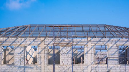 Concrete block wall with metal hip roof beam structure of incomplete modern office building in construction site against blue sky background
