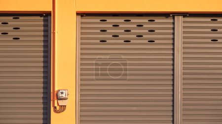 Gray roller shutter entrance door of yellow rental shophouse with electric meter and conduit pipe on concrete building wall, front view with copy space