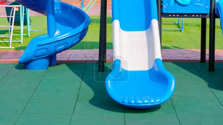 2 slides with playground equipment on green EPDM rubber mat floor with grass and stone tile floor in public outdoors playground area