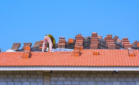 Construction worker is laying aluminum foil coated insulation and orange roof tiles on hip roof structure of modern house building against blue clear sky background