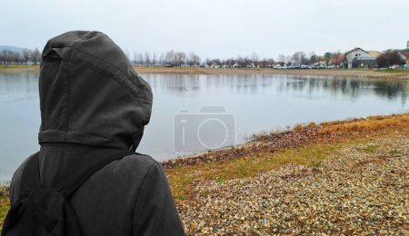 A girl in a dark suit with a hood on her head stands on the shore of the lake with her back to the camera