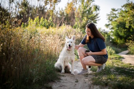 Téléchargez les photos : Une fille à lunettes caresse un chien blanc sur un chemin de terre dans la forêt. - en image libre de droit
