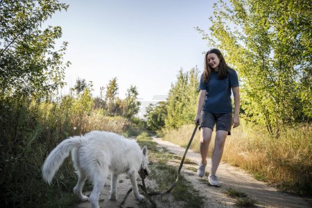 Téléchargez les photos : Une fille promène un chien blanc pendant la journée. Ils sont sur le fond de la forêt. - en image libre de droit