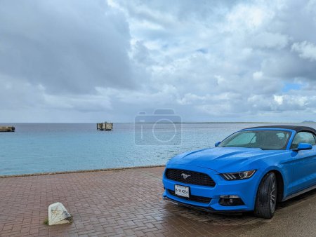Téléchargez les photos : Une Ford Mustang bleu vif se trouve sur le front de mer humide avec une vue sur la mer des Caraïbes verte et bleue et un nuage en arrière-plan à Kralendijk, Bonaire, Antilles sous le vent - en image libre de droit