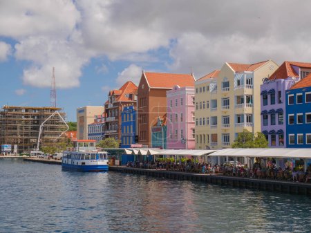 Téléchargez les photos : Bâtiments traditionnels et colorés de style hollandais sur le front de mer à Willemstad avec un bateau et des gens dans les cafés à quai. - en image libre de droit