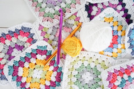 colorful woolen balls with multicolored granny squares and crochet hooks on white wooden ground handmade