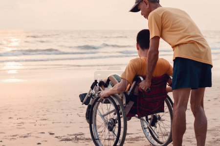 Foto de Un padre, cuidador o voluntario que ayuda a un adolescente a empujar la silla de ruedas a la playa al atardecer, hacer ejercicio y aprender sobre la naturaleza en el mar, viajar de vacaciones con la familia, concepto de salud mental. - Imagen libre de derechos