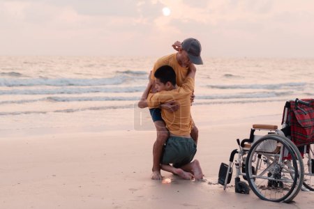 Foto de Un padre, cuidador o voluntario ayuda a mover al niño de la silla de ruedas para jugar a la arena en la playa del mar puesta del sol, momento hermoso, compartir, ministrar, cuidar, viajar de vacaciones con la familia, buen concepto de salud mental - Imagen libre de derechos