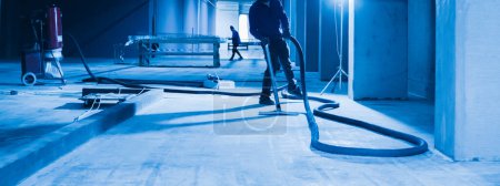 Téléchargez les photos : Les travailleurs utilisent des aspirateurs industriels pour nettoyer un sol en béton. L'image est dominée par des nuances de bleu, ajoutant à l'ambiance industrielle globale de la scène. - en image libre de droit
