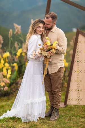 This beautiful photograph captures the tender embrace of a couple at their humanist wedding ceremony. The bride is holding a lovely bouquet of flowers while the backdrop features a stunning array of flowers, wood, and mountains.