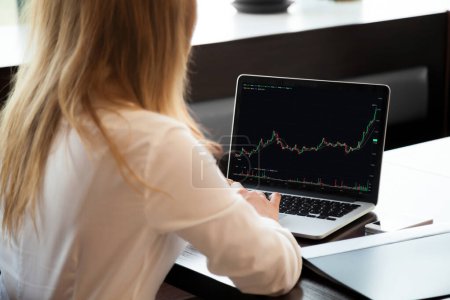 Unknown businesswoman trader makes stock market research, laptop screen view over female shoulder sit at desk analyzing graphs, charts showing good result, income and profit. Cryptocurrency, trading