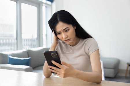 Foto de Mujer asiática joven preocupada usando teléfono inteligente en la mesa de casa, mirando la pantalla en pensamientos tristes y ansiosos, pensando, sintiéndose estresada, confundida, preocupada, tocando la cabeza, charlando - Imagen libre de derechos