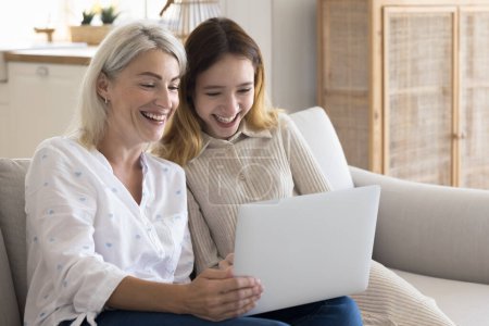 Photo for Cheerful mature mother spend time with teen daughter, sit on couch with laptop, watch funny video or movie, make purchase via web-store, learn new program, browse internet. Family leisure, technology - Royalty Free Image