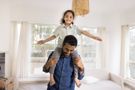 Photo for Cheerful young Indian father carrying cute little girl with open arms on shoulders, walking at home, smiling, laughing. Happy daddy and daughter playing airplane flight, enjoying family activity - Royalty Free Image