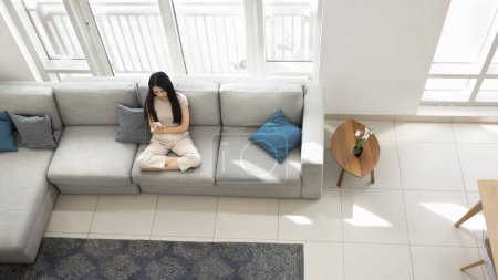 Photo for Young Asian woman using smart home application on mobile phone, sitting on large comfortable sofa, shopping with e-commerce app on smartphone, texting, browsing Internet. Banner shot, top view - Royalty Free Image