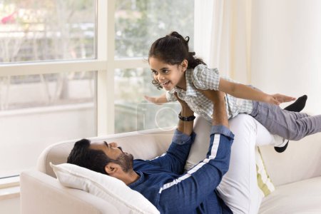 Téléchargez les photos : Joyeux jeune père indien couché sur le dos, tenant la petite fille sur les bras et les jambes, soulevant l'enfant dans les airs pour jouer à l'avion, rire, s'amuser, profiter des loisirs, la paternité, faire de l'acroyoga - en image libre de droit