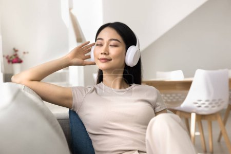 Téléchargez les photos : Joyeux rêveur jeune fille étudiante asiatique se détendre avec des airs ambiants préférés à la maison, écouter de la musique à partir d'écouteurs sans fil avec les yeux fermés, toucher les écouteurs, profiter de la détente - en image libre de droit