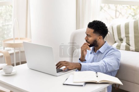 Foto de Preocupado joven freelancer africano pensando en el proyecto de negocios, la resolución de problemas, el trabajo en el ordenador portátil en casa. Estudiante serio estudiando en la computadora, mirando hacia otro lado, tocando la barbilla en pensamientos profundos - Imagen libre de derechos