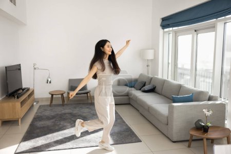 Foto de Feliz chica asiática joven adulta bailando en el acogedor interior de la sala de estar, divirtiéndose, disfrutando de la actividad en casa, entretenimiento, movimiento, celebrando el logro, el éxito. Disparo completo - Imagen libre de derechos