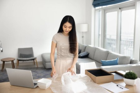 Positive young Asian customer woman unpacking parcel at home, opening package from Internet store, holding plastic wrap, packing fragile dish into paper box, standing at workplace table with laptop