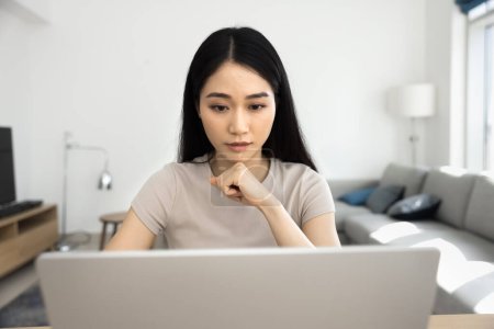 Téléchargez les photos : Focalisé jeune fille professionnelle asiatique sérieuse travaillant sur le projet d'affaires en ligne à la maison, assis à table avec ordinateur portable, penché menton sur la main, en utilisant la technologie Internet. Vue de face - en image libre de droit