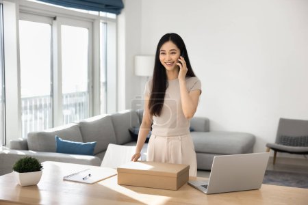 Happy young Asian consumer woman getting purchase from Internet store, holding paper cardboard box, talking on smartphone. Customer ordering delivery shipping service on mobile phone