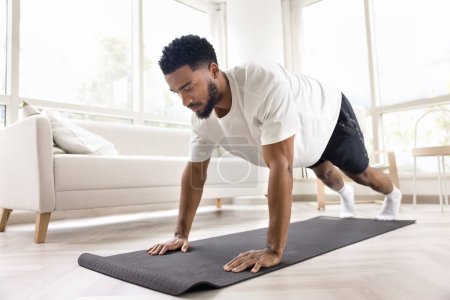 Téléchargez les photos : Focalisé athlétique fort homme africain faire des pompes, des exercices matinaux à la maison, prendre soin de la puissance, la force, le bien-être, le sport, le corps d'entraînement, en gardant la pose de planche statique sur le tapis de yoga - en image libre de droit