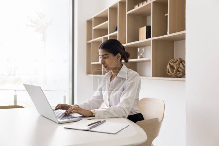 Photo for Serious young Indian businesswoman working at laptop in office white interior, typing on computer, using online technology, professional application for job communication - Royalty Free Image