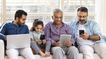 Photo for Online addicted Indian kid and men of four generations sitting together on sofa, using application, service, digital gadgets, holding laptop, smartphone, tablet, playing, browsing social media - Royalty Free Image