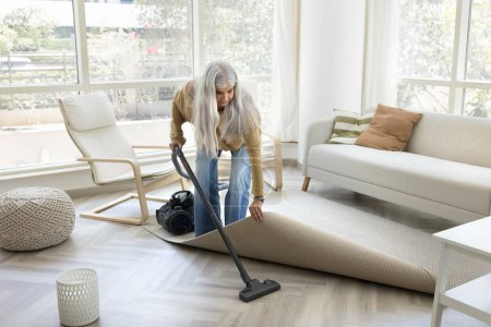Photo for Serious grey haired mature housemaid woman hoovering floor under rug, using vacuum cleaner, tidying up, doing household chores with electrical home appliance - Royalty Free Image