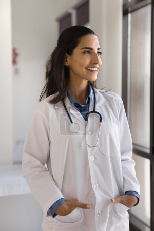 Happy confident young doctor woman posing at window in medical office, looking away with toothy smile, thinking on successful patients case. Positive medical practitioner woman vertical portrait