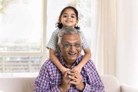 Téléchargez les photos : Joyeux jolie petite fille embrassant grand-père par derrière. Happy Indian grand-père piggybackking kid, regardant la caméra, souriant, profiter des loisirs avec l'enfant, posant pour le portrait de famille - en image libre de droit