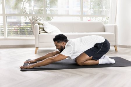 Téléchargez les photos : Heureux jeune homme afro-américain faire du yoga à la maison, garder la pose de lièvre, étirer les muscles du dos après l'exercice, corps d'entraînement sur le sol, profiter de l'activité à l'intérieur. Vue latérale - en image libre de droit