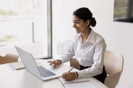 Photo for Happy young Indian office employee girl using laptop for Internet communication in office writing notes, smiling, talking on video call. Company intern watching business webinar - Royalty Free Image