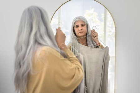 Foto de Elegante mujer hispana mayor de cabello gris natural que elige un suéter de gran tamaño para usar, cuidar el estilo, ropa de moda, mirarse en el espejo, probarse un paño en la percha en el vestuario - Imagen libre de derechos